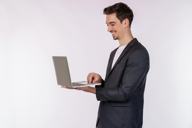 Retrato de jovem empresário sorridente e bonito segurando laptop nas mãos digitando e navegando em páginas da web isoladas em fundo branco