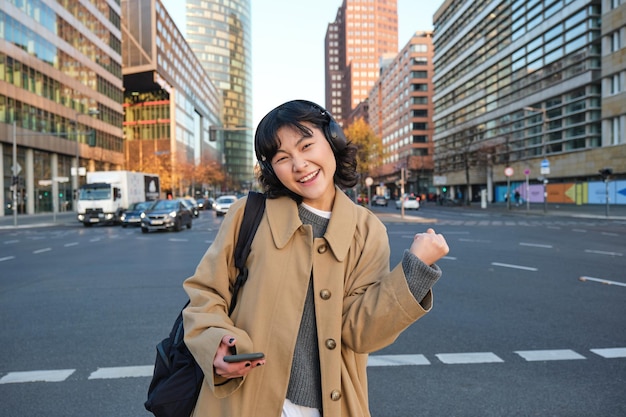 Retrato de jovem estudante asiática caminha na cidade, ouve música em fones de ouvido e usa telefone celular