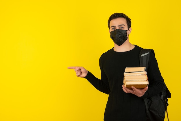 Retrato de jovem estudante com máscara médica, segurando livros da faculdade e apontando