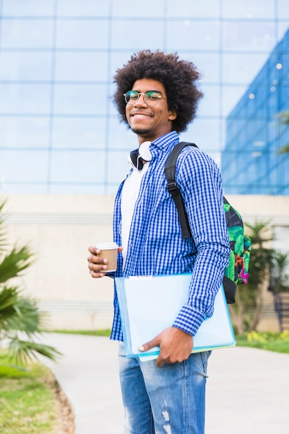 Retrato, de, jovem, estudante masculino, segurando, copo descartável, e, livros, em, mão, ficar, contra, campus