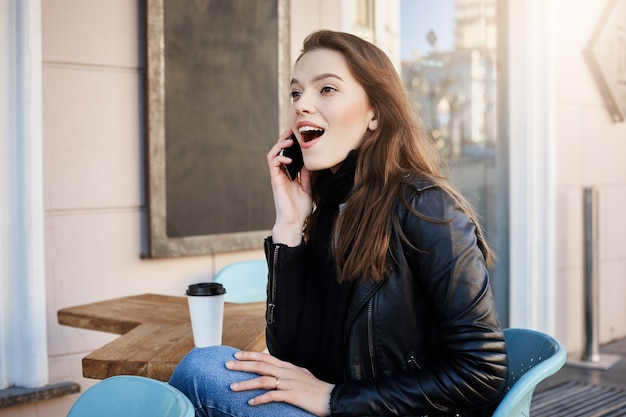 Retrato de jovem europeu feminino impressionado e animado com roupa elegante, sentado no café, tomando café e falando no smartphone