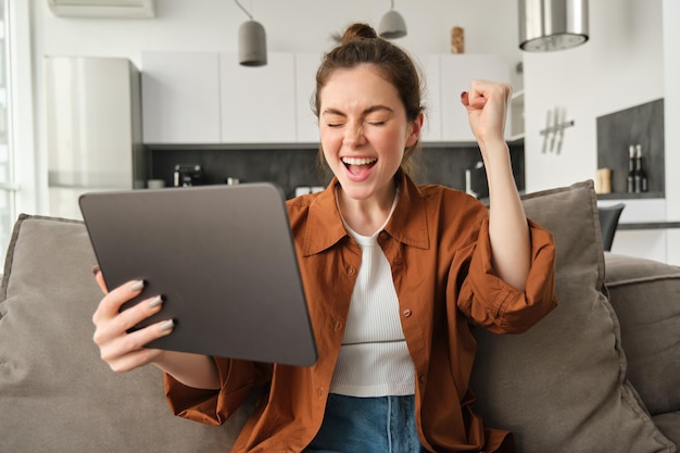 Retrato de jovem excitada com tablet digital sentada no sofá rindo e sorrindo vencendo em