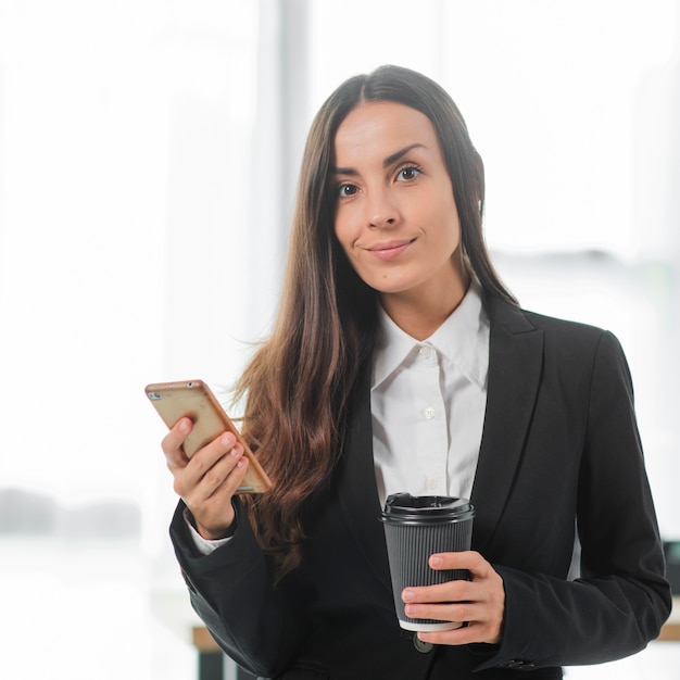 Retrato, de, jovem, executiva, segurando, smartphone, e, copo descartável, café, em, mãos