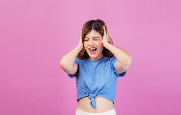 Retrato de jovem irritada com raiva vestindo camiseta casual cobrindo os ouvidos com as mãos e gritando em pé isolado sobre fundo rosa