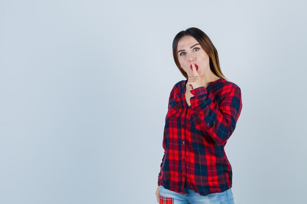 Retrato de jovem mostrando gesto de silêncio em uma camisa xadrez e olhando a misteriosa vista frontal