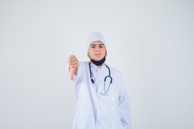 Retrato de jovem mostrando o polegar para baixo em uniforme branco, máscara e parecendo descontente