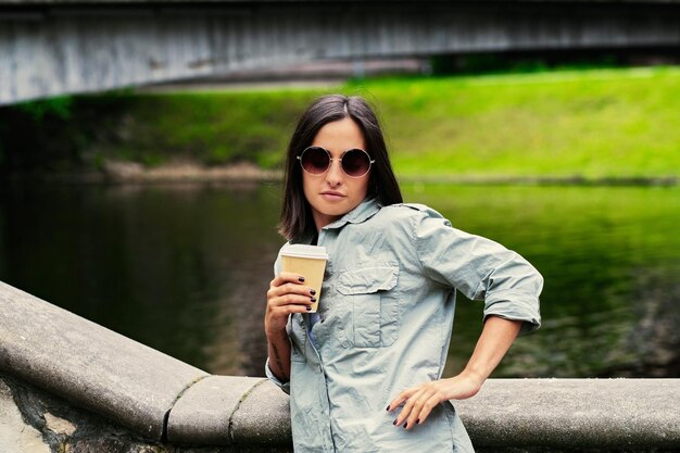 Retrato de jovem mulher atraente bebe café em movimento em um parque de verão perto do lago.