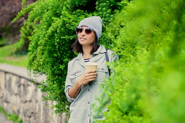 Retrato de jovem mulher atraente em óculos de sol detém xícara de café de papel em um parque verde de verão.