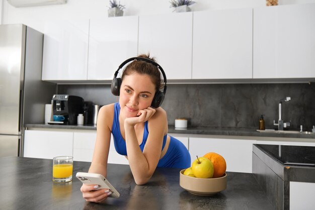 Foto grátis retrato de jovem mulher de fitness com fones de ouvido bebendo suco de laranja na cozinha e usando
