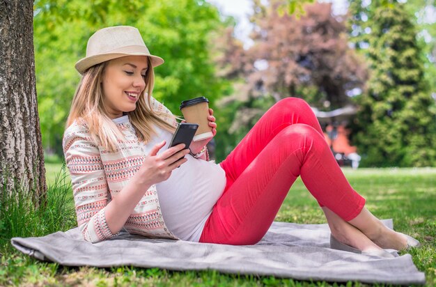 Retrato de jovem mulher feliz deitada na grama, segurando o telefone celular e café para ir. Retrato de corpo inteiro de mulher feliz deitado na grama, texting com telefone inteligente