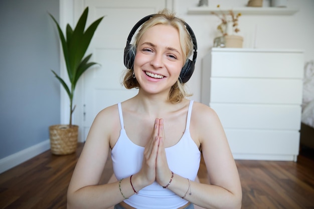 Retrato de jovem mulher relaxada sentada na sala com fones de ouvido, mãos juntas, meditando, ouvindo