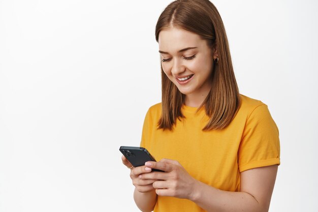 Retrato de jovem olha para a tela do smartphone e sorrindo, lendo mensagem no celular, usando aplicativo móvel ou mensageiro, em pé contra um fundo branco.