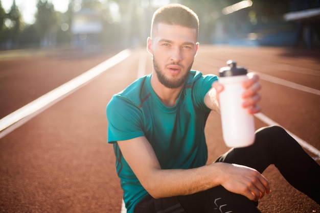 Foto grátis retrato de jovem olhando sonhadoramente na câmera mostrando garrafa esportiva branca enquanto passa o tempo na pista de corrida do estádio