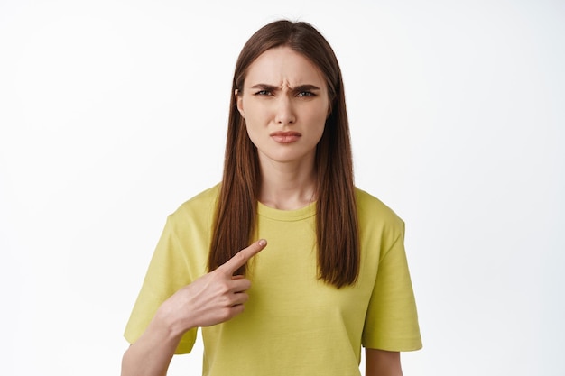 Retrato de jovem parecendo confuso enquanto aponta para si mesma, vestindo camiseta amarela casual, fundo branco