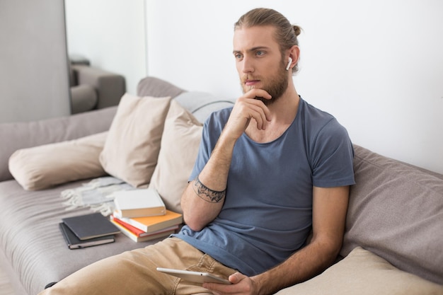 Retrato de jovem pensativo sentado no sofá cinza com fones de ouvido e o tablet nas mãos e sonhadoramente olhando de lado em casa