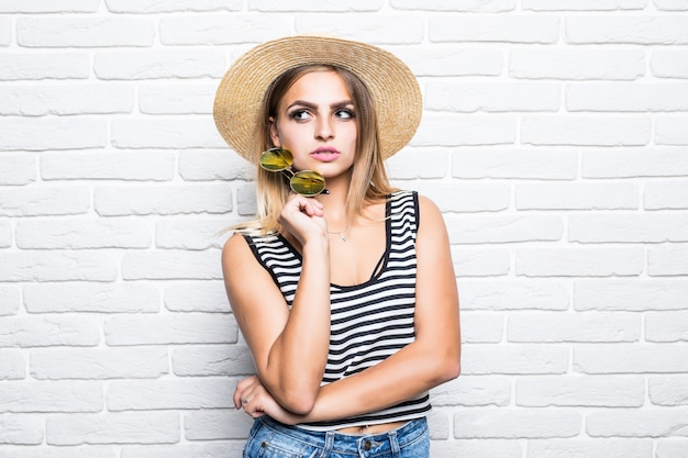 Retrato de jovem posando com chapéu de palha, aproveitando o verão em pé na parede de tijolos brancos