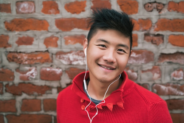 Retrato de jovem rapaz asiático ouvindo música com fones de ouvido ao ar livre contra a parede de tijolos. Conceito urbano.