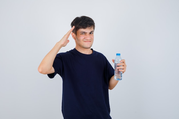 Retrato de jovem segurando uma garrafa de água, mostrando um gesto de saudação, franzindo os lábios enquanto franzia a testa em uma camiseta preta e parecendo confuso com a vista frontal