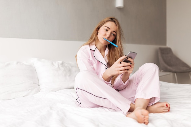 Retrato de jovem sentada na cama com escova de dentes enquanto estiver usando o celular em casa