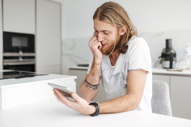 Retrato de jovem sentado com tablet digital na mão na cozinha em casa