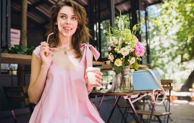 Retrato de jovem sorridente feliz mulher bonita sentada no café, bebendo café, roupa da moda para o verão, estilo hippie, vestido rosa de algodão, acessórios de vestuário da moda