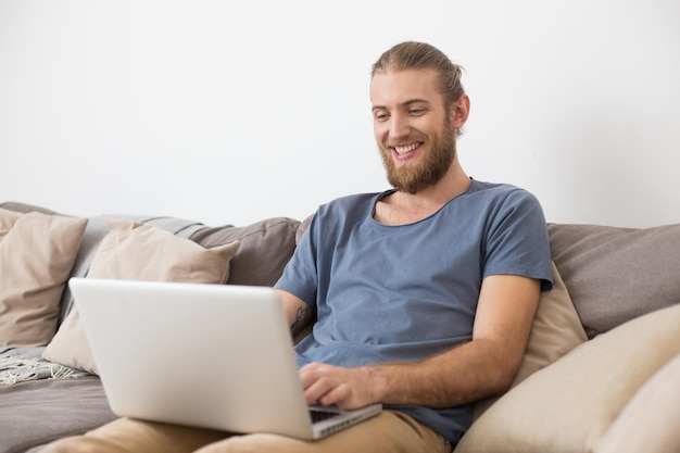 Retrato de jovem sorridente sentado no grande sofá cinza e trabalhando no laptop em casa