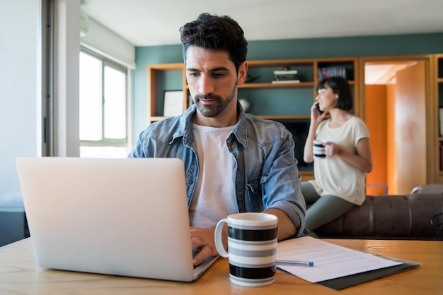 Retrato de jovem trabalhando com um laptop em casa enquanto uma mulher falando ao telefone no fundo