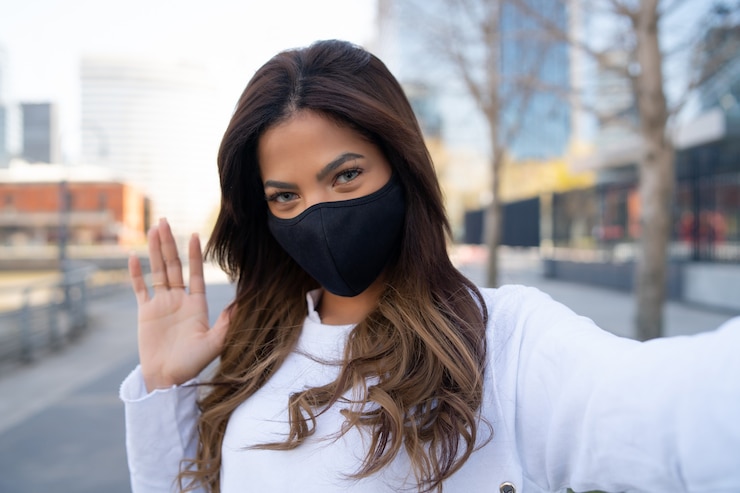 Foto grátis retrato de jovem usando máscara facial e tirando selfies enquanto acena a mão para dizer olá ao ar livre. conceito urbano. novo conceito de estilo de vida normal.