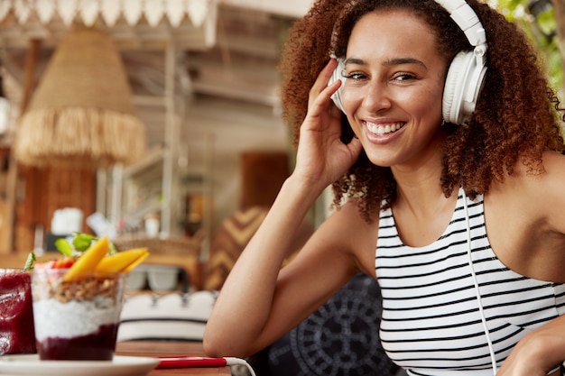 Retrato de lado de mulher morena com cabelo escuro usa fones de ouvido de alta qualidade e telefone celular para ouvir música ou livro de áudio, passa o tempo de lazer em um café, gosta de Internet de alta velocidade