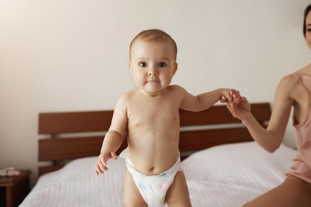 Retrato de lindo bebê recém-nascido em pé na cama na manhã mãe segurando a mão da criança. copie o espaço.