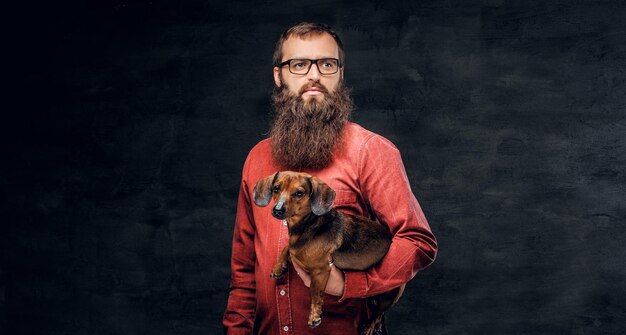 Retrato de macho barbudo de camisa vermelha detém um cão texugo marrom.