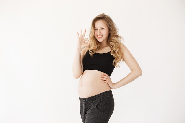 Foto grátis retrato de mãe grávida alegre bonita jovem com cabelos longos loiros, vestindo roupas pretas confortáveis, fazendo sinal de ok com a mão