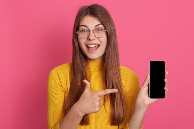 Retrato de magnético adorável muito feminino jovem abrindo a boca, fazendo a apresentação do novo smartphone, fazendo recomendações, segurando o dispositivo, fazendo a escolha, estar encantado. conceito de tecnologia.