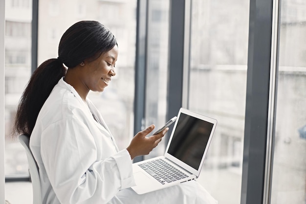 Retrato de médica negra sentada em seu escritório na clínica e usando um laptop