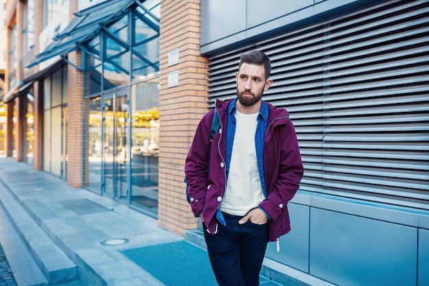 Foto grátis retrato de meia-lengua de um bonito homem barbudo vestido com roupas à moda de pé na rua contra o fundo da parede com área para conteúdo publicitário.