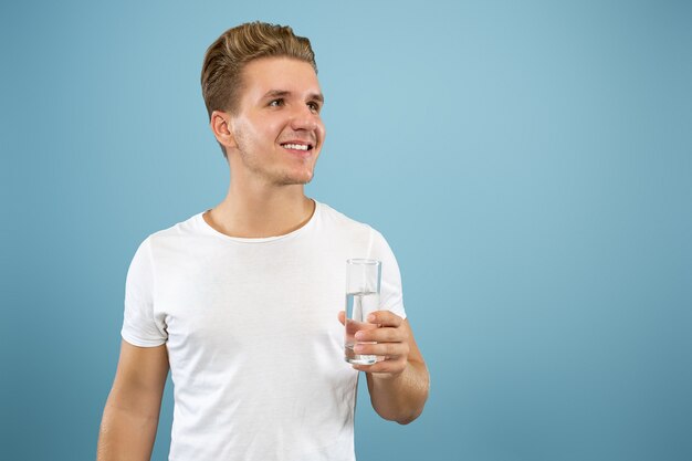 Retrato de meio comprimento de jovem caucasiano sobre fundo azul do estúdio. Lindo modelo masculino na camisa. Conceito de emoções humanas, expressão facial, vendas, anúncio. Gostando de beber água pura.