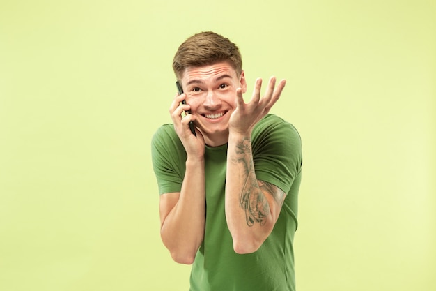Retrato de meio comprimento de jovem caucasiano sobre fundo verde do estúdio. lindo modelo masculino na camisa. conceito de emoções humanas, expressão facial, vendas, anúncio. falando no telefone e parece feliz.