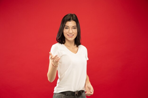 Retrato de meio comprimento de mulher jovem caucasiana em fundo vermelho studio. Bela modelo feminino em camisa branca. Conceito de emoções humanas, expressão facial. Mostrando algo, convidando, escolhendo.