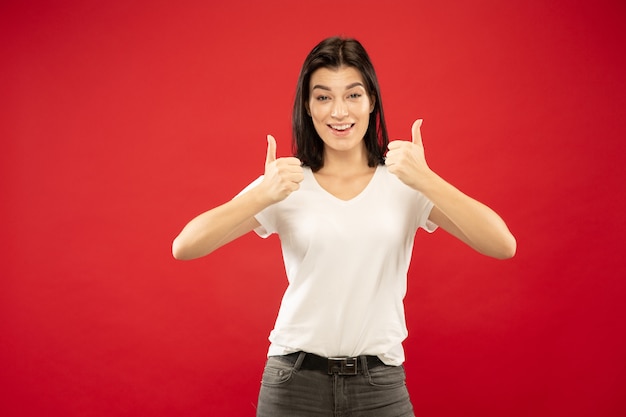 Retrato de meio comprimento de mulher jovem caucasiana em fundo vermelho studio. Bela modelo feminino em camisa branca. Conceito de emoções humanas, expressão facial. Mostrando o sinal de bom, ótimo.