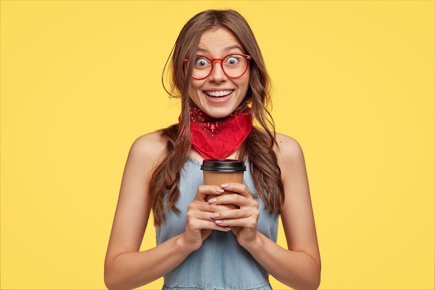 Retrato de menina alegre e alegre usa bandana vermelha, vestido jeans e óculos, segurando café para viagem em copo descartável de papel