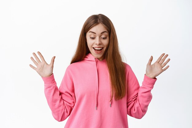 Retrato de menina animada ofegando e maravilhada, sorrindo fascinado, estendendo as mãos para os lados e olhando para baixo com uma expressão satisfeita, em pé sobre uma parede branca