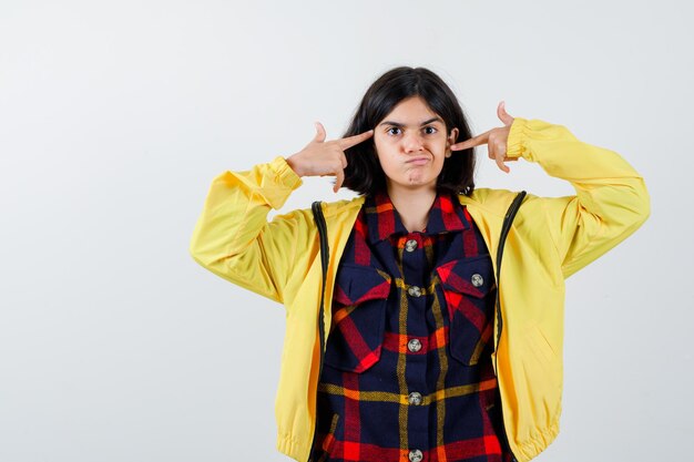 Retrato de menina apontando a cabeça com as mãos em uma camisa xadrez, jaqueta e olhando a vista frontal engraçada