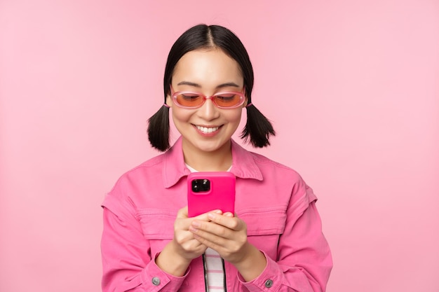 Retrato de menina asiática em óculos de sol usando smartphone Mulher olhando para celular navegando no aplicativo em cima de fundo rosa