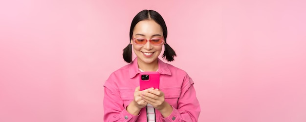 Retrato de menina asiática em óculos de sol usando smartphone Mulher olhando para celular navegando no aplicativo em cima de fundo rosa