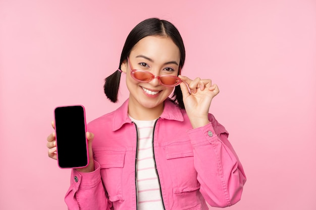 Retrato de menina asiática moderna elegante mostra interface de aplicativo de smartphone de tela de celular em óculos de sol contra fundo rosa