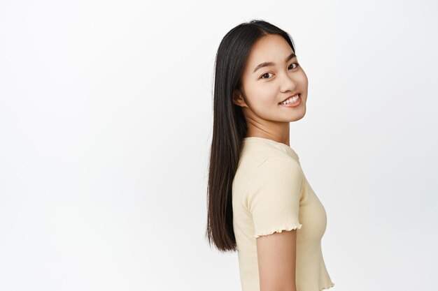 Retrato de menina asiática morena sorridente com cabelo saudável brilhante olhando para a câmera confiante e feliz em pé sobre fundo branco
