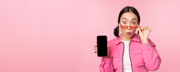 Retrato de menina asiática mostrando a tela do celular reagindo surpreso em pé sobre fundo rosa