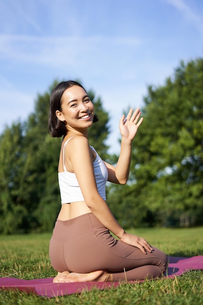 Retrato de menina asiática na aula de fitness no parque sentado no tapete de borracha e acenando com a mão para a câmera diz que ele