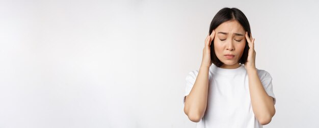 Retrato de menina asiática sentindo enxaqueca de dor de cabeça ou estando doente em camiseta branca sobre ba branco