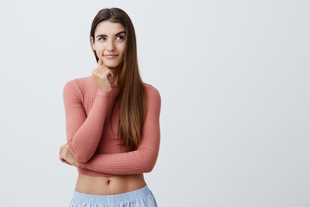 Retrato de menina atraente estudante caucasiano sexy com longos cabelos escuros em roupas elegantes, segurando o queixo com e, olhando de lado com a expressão do rosto sexy